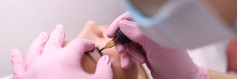 A technician in pink gloves using a precision tattoo needle to apply permanent eyeliner to a client's upper lash line